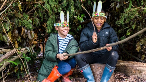 2 children sitting on a log wearing paper bunny ears and playing with sticks